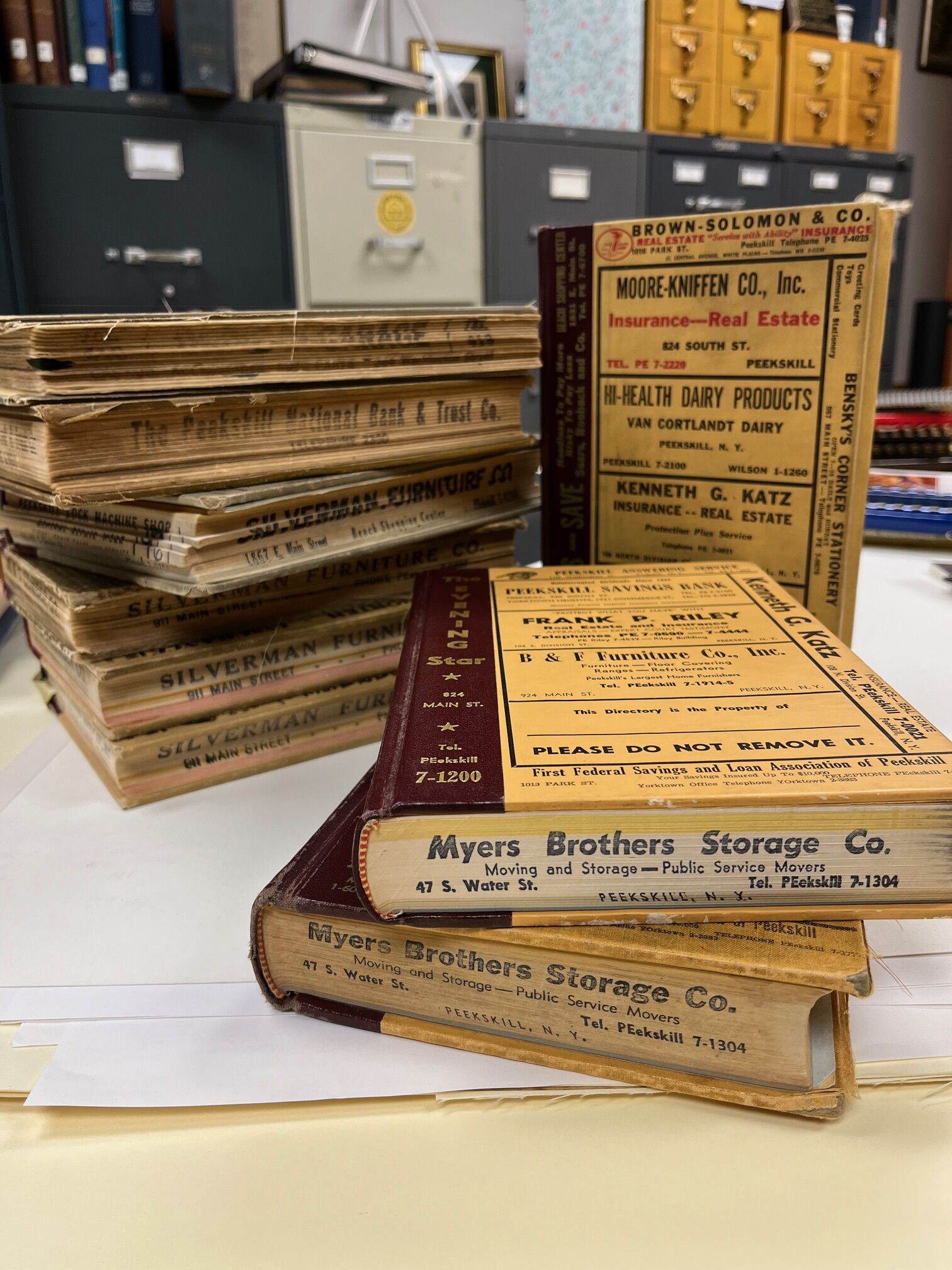Stack of city directories with burgundy binding and yellow covers in the foreground of picture with “Myers brothers Storage Co,” Background features filing cabinet